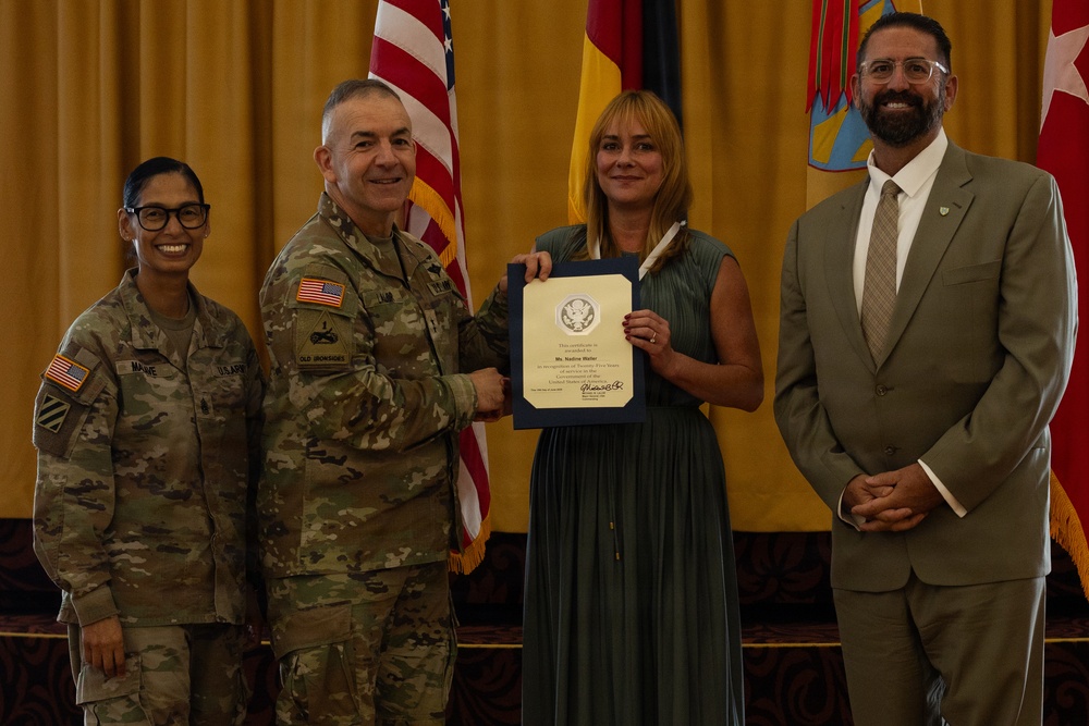 A formal military ceremony in which a U.S. Army officer presents a certificate to a civilian woman, flanked by another soldier and a civilian man in a suit. The setting appears to be an official awards or recognition event, with American and German flags visible in the background. The certificate appears to recognize years of service to the United States government.