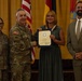 A formal military ceremony in which a U.S. Army officer presents a certificate to a civilian woman, flanked by another soldier and a civilian man in a suit. The setting appears to be an official awards or recognition event, with American and German flags visible in the background. The certificate appears to recognize years of service to the United States government.