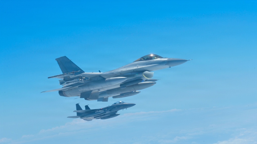 Two military fighter jets are flying in formation against a clear blue sky. The foreground aircraft is a General Dynamics F-16 Fighting Falcon, identifiable by its distinctive bubble canopy, single engine, and blended wing-body design. A second aircraft, appearing to be an F-15 Eagle, flies in loose formation below and behind the lead aircraft.
