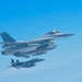 Two military fighter jets are flying in formation against a clear blue sky. The foreground aircraft is a General Dynamics F-16 Fighting Falcon, identifiable by its distinctive bubble canopy, single engine, and blended wing-body design. A second aircraft, appearing to be an F-15 Eagle, flies in loose formation below and behind the lead aircraft.