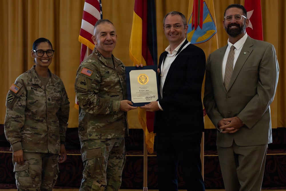 A formal military ceremony is taking place in which a U.S. Army officer is presenting a certificate or award to a civilian male. Four individuals are posed together on a stage with flags in the background, including the U.S. flag and what appears to be a German flag. The setting suggests an official recognition or commendation event.
