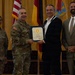 A formal military ceremony is taking place in which a U.S. Army officer is presenting a certificate or award to a civilian male. Four individuals are posed together on a stage with flags in the background, including the U.S. flag and what appears to be a German flag. The setting suggests an official recognition or commendation event.