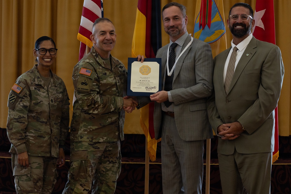 A formal military ceremony is taking place in which a uniformed Army officer is presenting a certificate or award to a civilian man in a gray suit. The recipient is wearing a medallion or lanyard around his neck, suggesting a formal recognition event. Four individuals are posed together in front of multiple flags, including the U.S. flag and military unit colors.