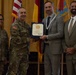 A formal military ceremony is taking place in which a uniformed Army officer is presenting a certificate or award to a civilian man in a gray suit. The recipient is wearing a medallion or lanyard around his neck, suggesting a formal recognition event. Four individuals are posed together in front of multiple flags, including the U.S. flag and military unit colors.