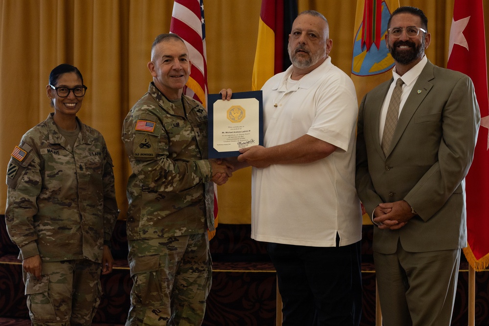 A military award or recognition ceremony is taking place indoors, with a uniformed U.S. Army officer presenting a framed certificate to a civilian man in a white polo shirt. Two additional individuals — a female soldier in Army Combat Uniform and a civilian man in a tan suit — stand alongside as witnesses. Multiple flags, including the U.S. flag and military unit flags, are visible in the background.