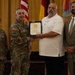 A military award or recognition ceremony is taking place indoors, with a uniformed U.S. Army officer presenting a framed certificate to a civilian man in a white polo shirt. Two additional individuals — a female soldier in Army Combat Uniform and a civilian man in a tan suit — stand alongside as witnesses. Multiple flags, including the U.S. flag and military unit flags, are visible in the background.