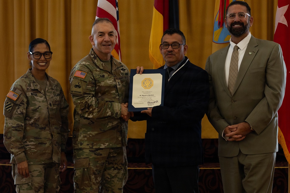 A formal military ceremony in which a U.S. Army officer is presenting a certificate to a civilian recipient. Four individuals are posed together: two in U.S. Army combat uniforms, one civilian in a dark suit holding the certificate, and one civilian in a tan suit. The setting appears to be an official venue with U.S. and military flags in the background.
