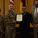 A formal military ceremony in which a U.S. Army officer is presenting a certificate to a civilian recipient. Four individuals are posed together: two in U.S. Army combat uniforms, one civilian in a dark suit holding the certificate, and one civilian in a tan suit. The setting appears to be an official venue with U.S. and military flags in the background.
