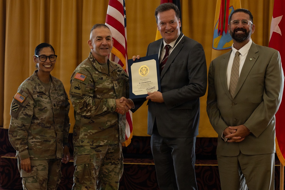 A formal military award ceremony is taking place, with a U.S. Army officer presenting a framed certificate to a civilian man in a gray suit. Two additional individuals — a female soldier and a civilian man in a tan suit — stand alongside as witnesses. The setting appears to be an official military venue with American and unit flags displayed in the background.