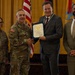 A formal military award ceremony is taking place, with a U.S. Army officer presenting a framed certificate to a civilian man in a gray suit. Two additional individuals — a female soldier and a civilian man in a tan suit — stand alongside as witnesses. The setting appears to be an official military venue with American and unit flags displayed in the background.