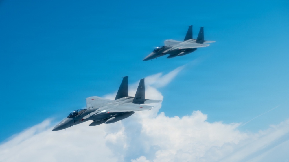 Two military fighter jets, identifiable as F-15 Eagles, are flying in formation against a bright blue sky with scattered clouds below. The foreground aircraft is generating visible condensation vapor trails along its fuselage, likely due to high humidity and aerodynamic pressure changes. Both aircraft appear to be in a coordinated flight pattern, photographed from a nearby aircraft.