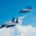 Two military fighter jets, identifiable as F-15 Eagles, are flying in formation against a bright blue sky with scattered clouds below. The foreground aircraft is generating visible condensation vapor trails along its fuselage, likely due to high humidity and aerodynamic pressure changes. Both aircraft appear to be in a coordinated flight pattern, photographed from a nearby aircraft.