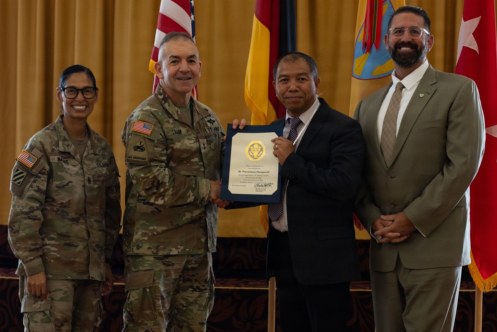 A formal military recognition ceremony where a civilian employee is receiving a certificate of appreciation, likely for years of service. Two U.S. Army soldiers in MultiCam/OCP uniforms flank a civilian recipient holding a blue-framed certificate, with another civilian in a suit standing to the right. The setting appears to be an official military venue with multiple flags displayed in the background.
