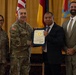 A formal military recognition ceremony where a civilian employee is receiving a certificate of appreciation, likely for years of service. Two U.S. Army soldiers in MultiCam/OCP uniforms flank a civilian recipient holding a blue-framed certificate, with another civilian in a suit standing to the right. The setting appears to be an official military venue with multiple flags displayed in the background.