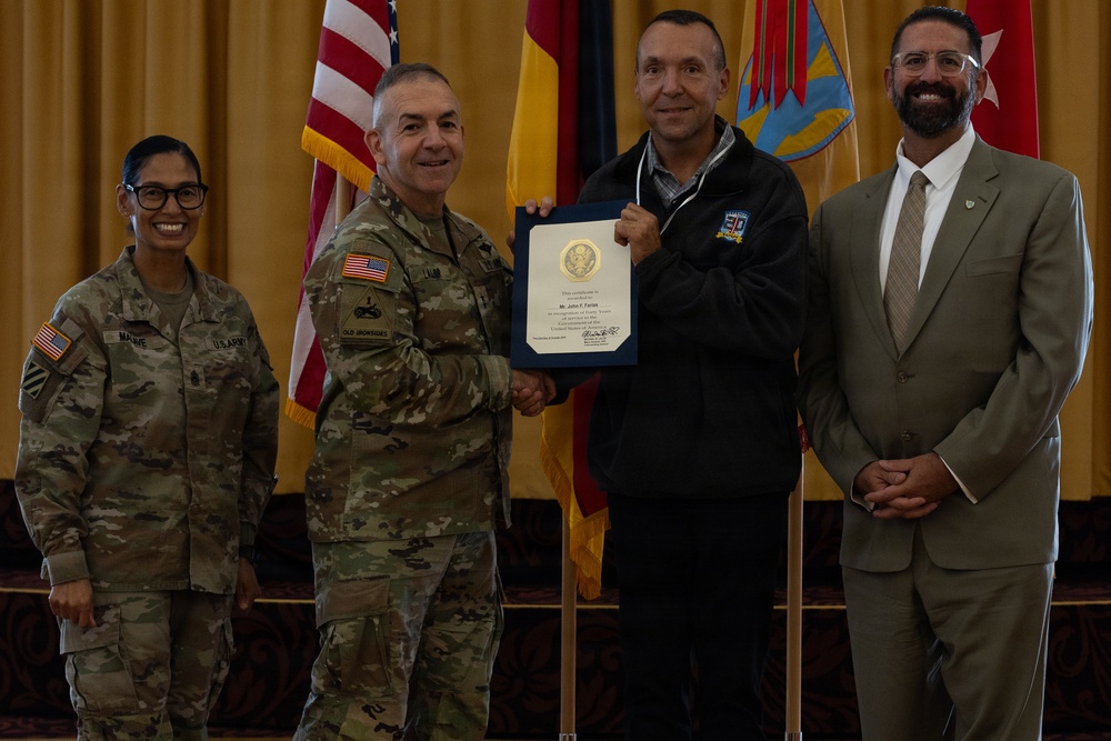 A military award ceremony is taking place in a formal indoor setting. A U.S. Army officer in camouflage uniform is presenting a certificate to a civilian man, while two other individuals — a female soldier and a civilian man in a suit — stand alongside them. The backdrop features multiple flags including the U.S. flag and what appears to be unit or state flags.