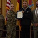 A military award ceremony is taking place in a formal indoor setting. A U.S. Army officer in camouflage uniform is presenting a certificate to a civilian man, while two other individuals — a female soldier and a civilian man in a suit — stand alongside them. The backdrop features multiple flags including the U.S. flag and what appears to be unit or state flags.