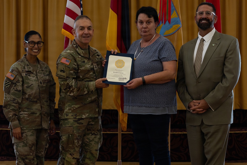 A military award ceremony is taking place in what appears to be a formal venue. A uniformed Army officer is presenting a certificate or award to a civilian woman, while two other individuals — a female soldier and a civilian man in a suit — stand alongside them. Flags including the American flag and the German flag are visible in the background.