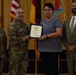 A military award ceremony is taking place in what appears to be a formal venue. A uniformed Army officer is presenting a certificate or award to a civilian woman, while two other individuals — a female soldier and a civilian man in a suit — stand alongside them. Flags including the American flag and the German flag are visible in the background.