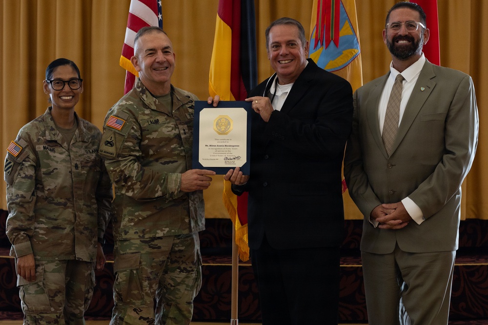 A formal military ceremony in which a uniformed Army officer presents a certificate to a civilian recipient. Four individuals are posed together for a photo, with two in military camouflage uniforms and two in civilian attire. The setting appears to be an official venue with flags including the U.S. flag and what appears to be a New Mexico state flag in the background.