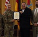 A formal military ceremony in which a uniformed Army officer presents a certificate to a civilian recipient. Four individuals are posed together for a photo, with two in military camouflage uniforms and two in civilian attire. The setting appears to be an official venue with flags including the U.S. flag and what appears to be a New Mexico state flag in the background.
