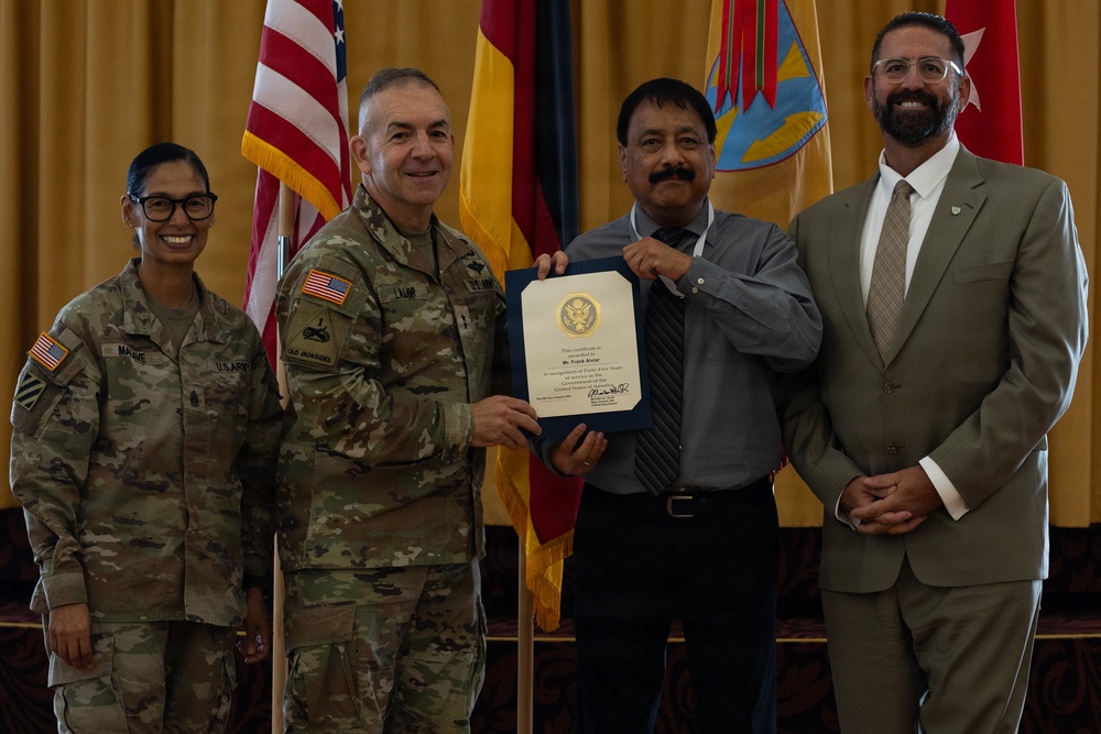 A formal military ceremony in which a U.S. Army officer presents a certificate or award to a civilian recipient. Four individuals are posed together: two in U.S. Army combat uniforms, one civilian recipient holding the certificate, and one civilian in a suit. Multiple flags are displayed in the background, indicating an official military installation setting.