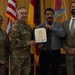 A formal military ceremony in which a U.S. Army officer presents a certificate or award to a civilian recipient. Four individuals are posed together: two in U.S. Army combat uniforms, one civilian recipient holding the certificate, and one civilian in a suit. Multiple flags are displayed in the background, indicating an official military installation setting.