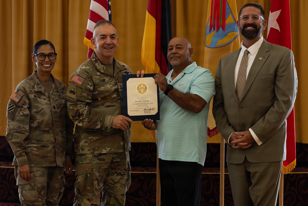 A formal military ceremony in which a uniformed Army officer presents a certificate to a civilian man. Three other individuals are present, including a female soldier and a man in a business suit. The group is posed in front of multiple flags including the U.S. flag and military unit colors.