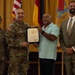 A formal military ceremony in which a uniformed Army officer presents a certificate to a civilian man. Three other individuals are present, including a female soldier and a man in a business suit. The group is posed in front of multiple flags including the U.S. flag and military unit colors.