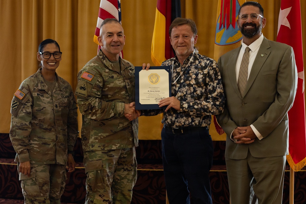 A formal military ceremony in which a uniformed U.S. Army officer is presenting a certificate or commendation to a civilian recipient. Four individuals are posed together in front of a display of flags including the U.S. flag, German flag, and what appears to be a unit or state flag. The setting appears to be an official awards or recognition event held indoors.
