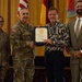 A formal military ceremony in which a uniformed U.S. Army officer is presenting a certificate or commendation to a civilian recipient. Four individuals are posed together in front of a display of flags including the U.S. flag, German flag, and what appears to be a unit or state flag. The setting appears to be an official awards or recognition event held indoors.
