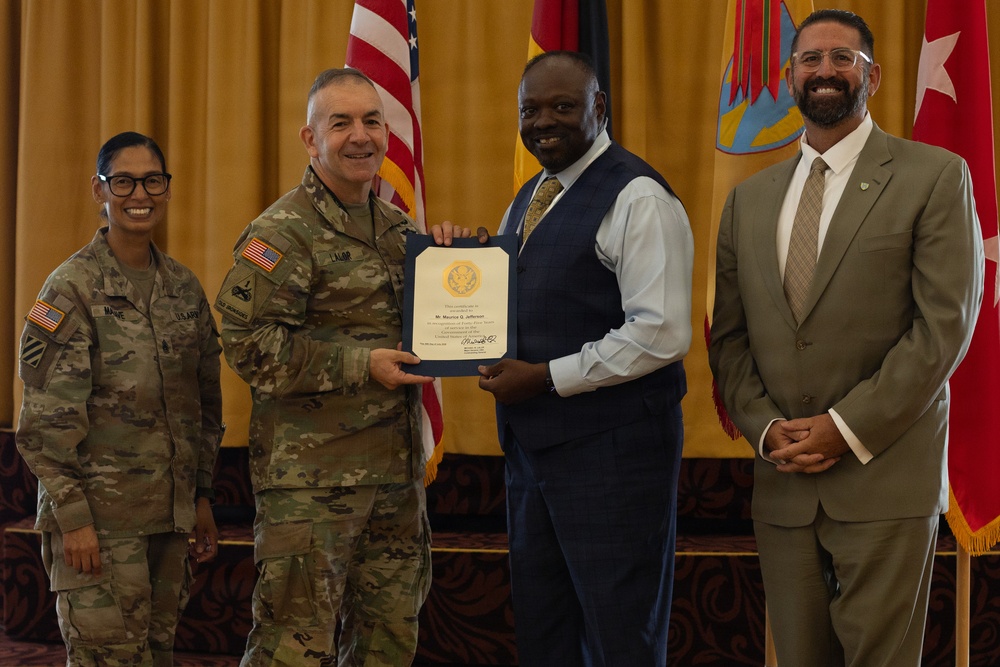 A formal military ceremony in which a uniformed Army officer is presenting a certificate or award to a civilian man in a blue suit. Four individuals are posed together for a photo, including two military personnel in Army Combat Uniforms and two civilians. The setting appears to be an official military venue with American and unit flags visible in the background.