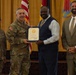 A formal military ceremony in which a uniformed Army officer is presenting a certificate or award to a civilian man in a blue suit. Four individuals are posed together for a photo, including two military personnel in Army Combat Uniforms and two civilians. The setting appears to be an official military venue with American and unit flags visible in the background.