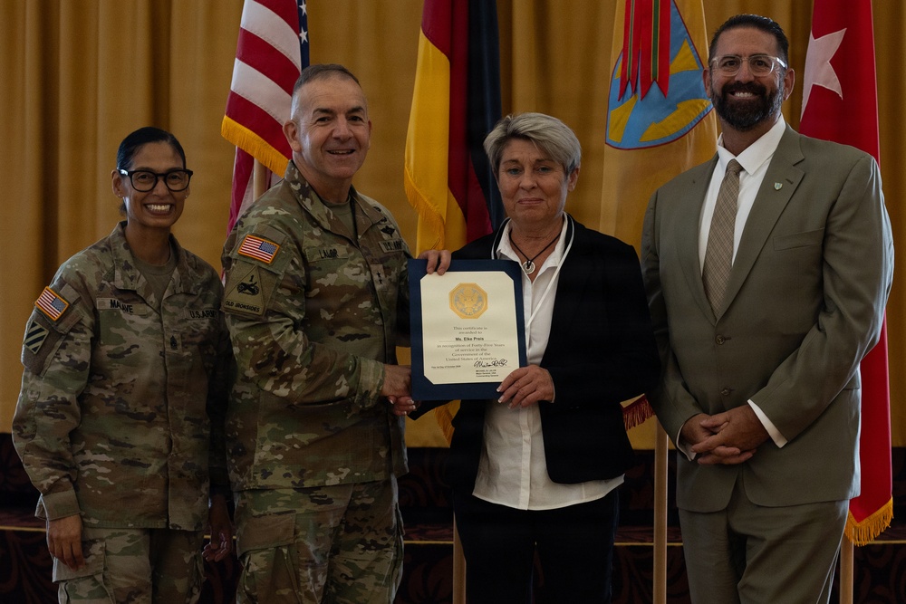 A formal military recognition ceremony in which a certificate is being presented to a civilian woman by a U.S. Army officer. Four individuals are posed together: two uniformed soldiers, one civilian woman receiving the award, and one civilian man in a suit. Multiple flags, including the U.S. flag and what appears to be a German flag, are visible in the background.