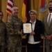 A formal military recognition ceremony in which a certificate is being presented to a civilian woman by a U.S. Army officer. Four individuals are posed together: two uniformed soldiers, one civilian woman receiving the award, and one civilian man in a suit. Multiple flags, including the U.S. flag and what appears to be a German flag, are visible in the background.