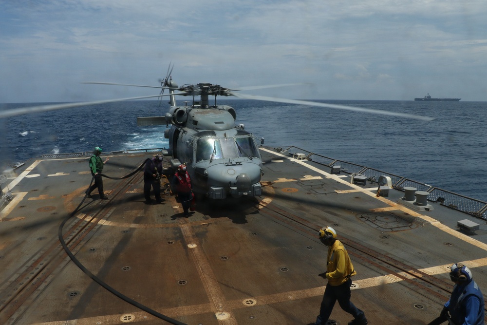 A military helicopter, consistent with an SH-60 Seahawk variant, is on the flight deck of a naval vessel with rotors spinning. Deck crew members in color-coded jerseys are conducting what appears to be a hot refueling (HIFR) or servicing operation, with a fuel hose visible. A second large naval vessel, possibly an aircraft carrier or amphibious assault ship, is visible on the horizon in the background.