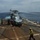 A military helicopter, consistent with an SH-60 Seahawk variant, is on the flight deck of a naval vessel with rotors spinning. Deck crew members in color-coded jerseys are conducting what appears to be a hot refueling (HIFR) or servicing operation, with a fuel hose visible. A second large naval vessel, possibly an aircraft carrier or amphibious assault ship, is visible on the horizon in the background.