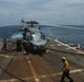 A military helicopter, consistent with an SH-60 Seahawk variant, is on the flight deck of a naval vessel with rotors spinning. Deck crew members in color-coded jerseys are conducting what appears to be a hot refueling (HIFR) or servicing operation, with a fuel hose visible. A second large naval vessel, possibly an aircraft carrier or amphibious assault ship, is visible on the horizon in the background.