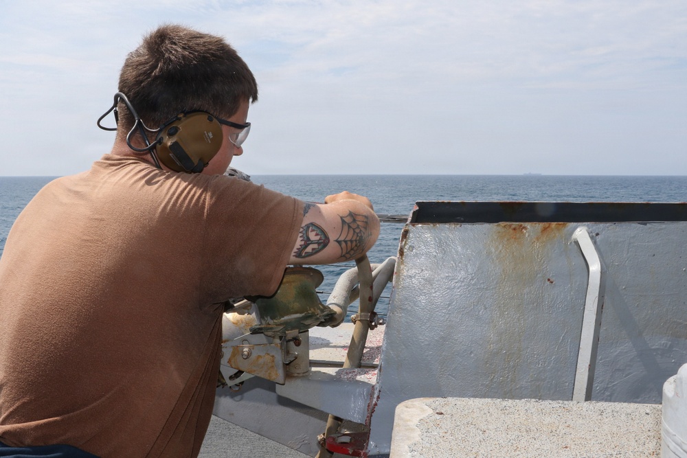 A person wearing hearing protection and safety glasses is positioned at what appears to be a weapon mount or observation post on the deck of a naval vessel. They are looking out toward the open ocean, leaning over a metal shield or gun mount. A tattoo featuring a spiderweb design is visible on their right arm.