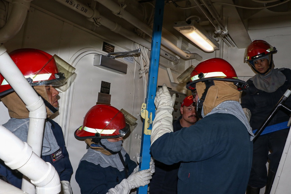 A group of five personnel wearing red safety helmets and protective gear are working together inside what appears to be a naval vessel or industrial facility. They are handling a blue metal pole or shoring device near a watertight door, suggesting damage control or emergency response training. The confined interior space features white-painted bulkheads, overhead piping, and fluorescent lighting typical of a military ship.