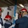 A group of five personnel wearing red safety helmets and protective gear are working together inside what appears to be a naval vessel or industrial facility. They are handling a blue metal pole or shoring device near a watertight door, suggesting damage control or emergency response training. The confined interior space features white-painted bulkheads, overhead piping, and fluorescent lighting typical of a military ship.