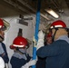 A group of five personnel wearing red safety helmets and protective gear are working together inside what appears to be a naval vessel or industrial facility. They are handling a blue metal pole or shoring device near a watertight door, suggesting damage control or emergency response training. The confined interior space features white-painted bulkheads, overhead piping, and fluorescent lighting typical of a military ship.