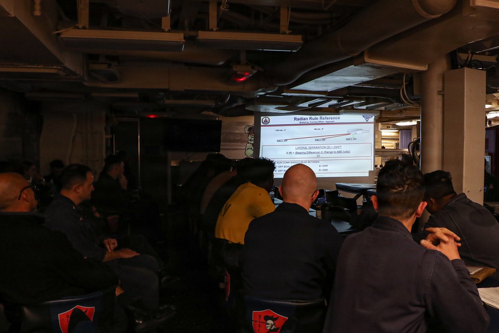 A group of military personnel are attending a briefing or training session in what appears to be a shipboard compartment. They are seated facing a projected presentation screen displaying a 'Radian Rule Reference' slide, which contains navigational or tactical formulas and diagrams. The setting, chair markings, and uniforms suggest a U.S. Navy vessel.