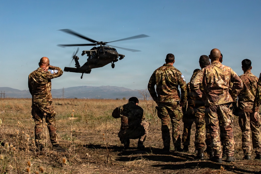 A group of military personnel in camouflage uniforms observe a UH-60 Black Hawk helicopter hovering at low altitude over an open field. Several soldiers are shielding their eyes or crouching due to rotor wash and debris. The soldiers appear to wear UN (United Nations) insignia patches on their shoulders, suggesting a peacekeeping or multinational training context.