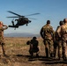 A group of military personnel in camouflage uniforms observe a UH-60 Black Hawk helicopter hovering at low altitude over an open field. Several soldiers are shielding their eyes or crouching due to rotor wash and debris. The soldiers appear to wear UN (United Nations) insignia patches on their shoulders, suggesting a peacekeeping or multinational training context.