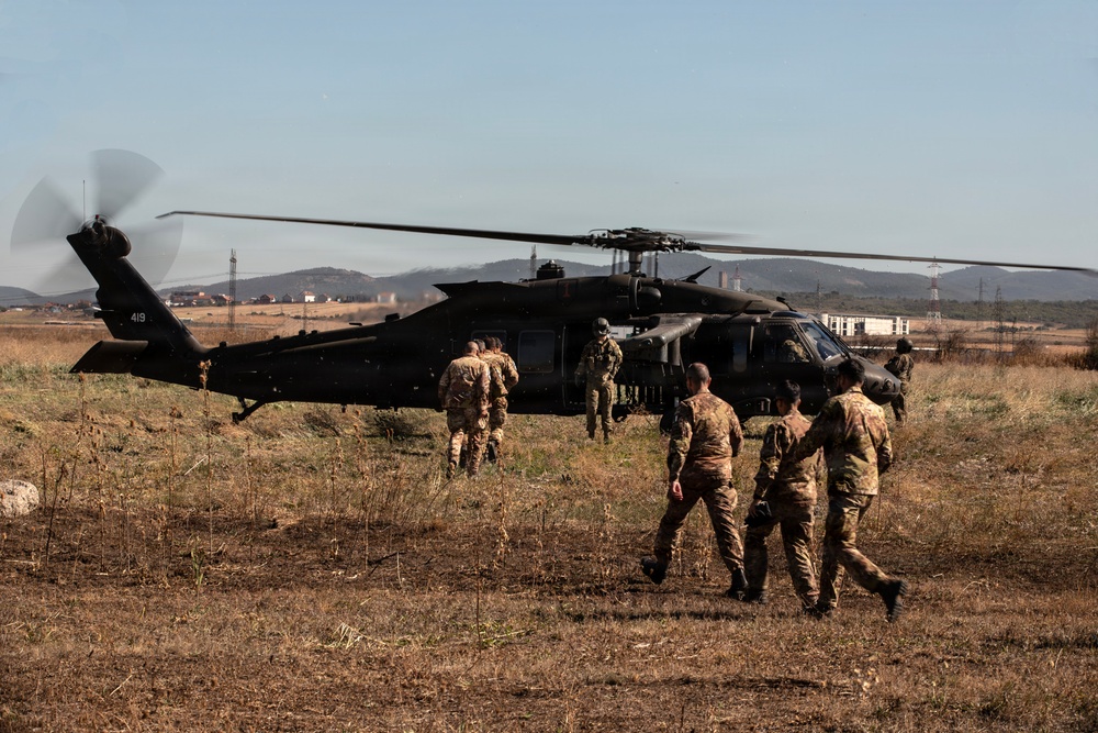 A group of military personnel in camouflage uniforms are approaching or boarding a black military helicopter, identified by the tail number '419', on an open grassy field. The helicopter's rotors are spinning, indicating it is either preparing for takeoff or has recently landed. The setting appears to be a semi-rural area with hills, buildings, and power lines visible in the background.