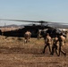 A group of military personnel in camouflage uniforms are approaching or boarding a black military helicopter, identified by the tail number '419', on an open grassy field. The helicopter's rotors are spinning, indicating it is either preparing for takeoff or has recently landed. The setting appears to be a semi-rural area with hills, buildings, and power lines visible in the background.