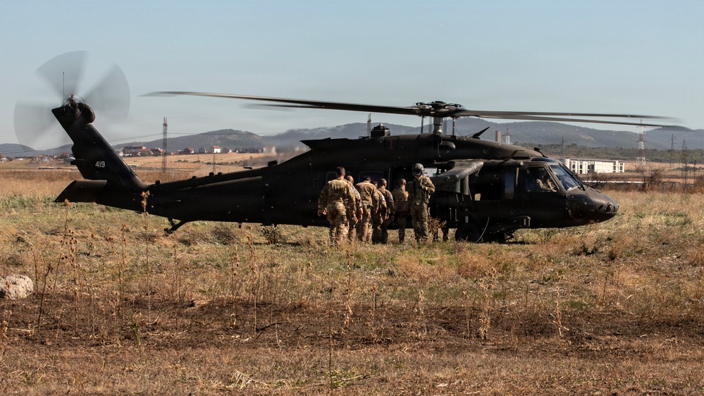 A black military helicopter, identifiable as a UH-60 Black Hawk with tail number 419, is landed on an open grassy field with rotors spinning. A group of soldiers in camouflage uniforms are boarding or staging near the aircraft's side door. The background shows a semi-rural landscape with hills, power transmission towers, and scattered buildings under a clear sky.