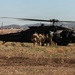 A black military helicopter, identifiable as a UH-60 Black Hawk with tail number 419, is landed on an open grassy field with rotors spinning. A group of soldiers in camouflage uniforms are boarding or staging near the aircraft's side door. The background shows a semi-rural landscape with hills, power transmission towers, and scattered buildings under a clear sky.