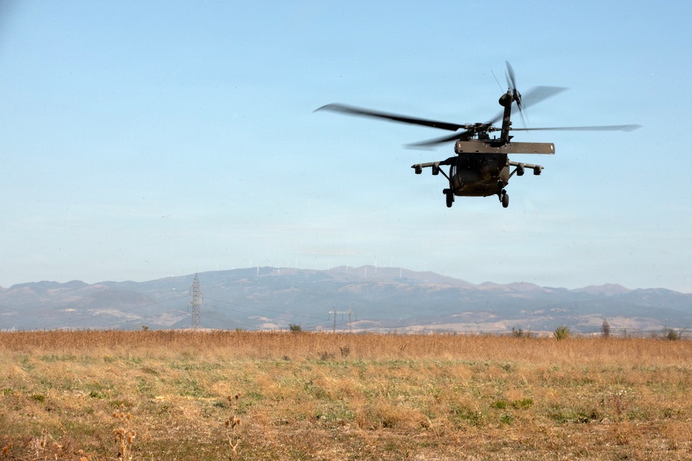 A military helicopter, consistent with a UH-60 Black Hawk, is captured in low-altitude flight over an open, dry grassland field. The aircraft appears to be either landing or taking off, with rotor blades visibly blurred due to rotation speed. The background features a mountain range, power transmission towers, and what appear to be wind turbines on distant ridgelines.