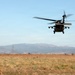 A military helicopter, consistent with a UH-60 Black Hawk, is captured in low-altitude flight over an open, dry grassland field. The aircraft appears to be either landing or taking off, with rotor blades visibly blurred due to rotation speed. The background features a mountain range, power transmission towers, and what appear to be wind turbines on distant ridgelines.
